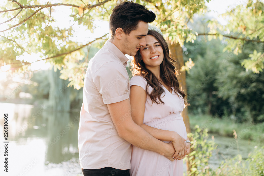 Fototapeta premium Man hugs beautiful pregnant woman tender standing before the lake in the rays of evening sun