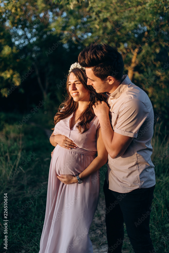 Man hugs beautiful pregnant woman tender standing before the lake in the rays of evening sun Man hugs beautiful pregnant woman tender standing before the lake in the rays of evening sun