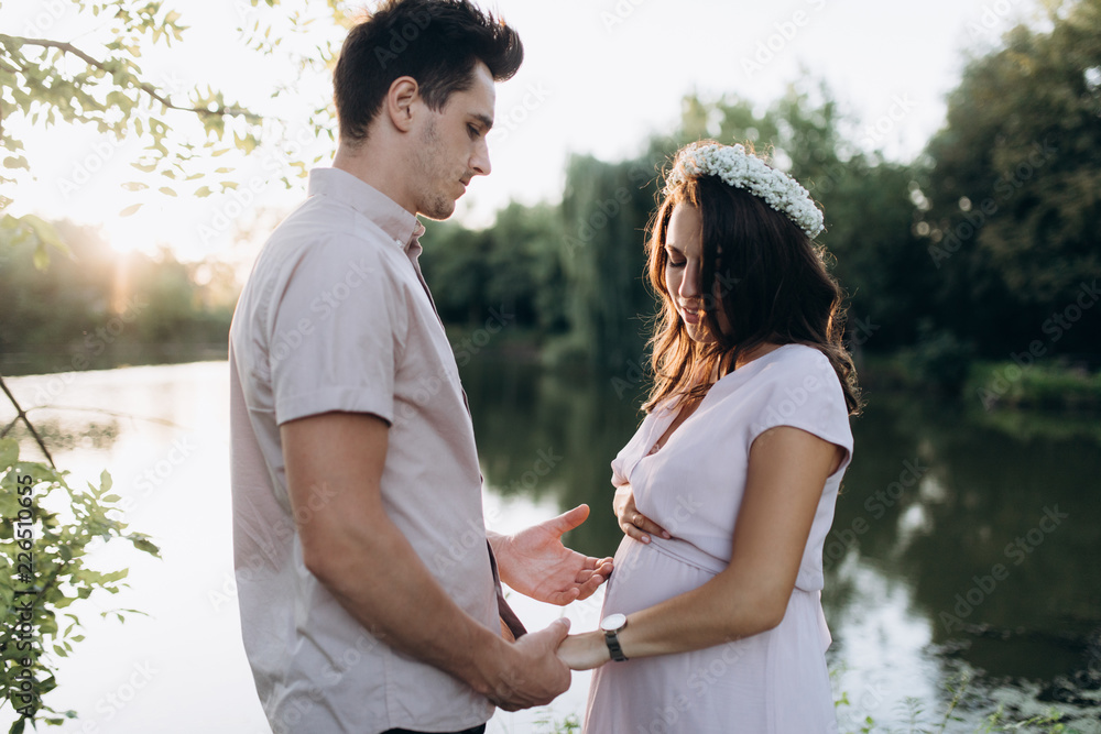 Man hugs beautiful pregnant woman tender standing before the lake in the rays of evening sun