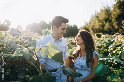 Cheerful man and pregnant woman hug each other tender standing in the field with tall sunflowers around them © kristina_1994