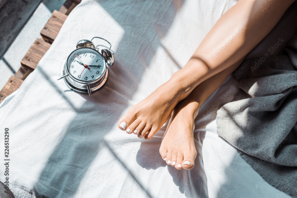 Obraz premium partial top view of female feet and alarm clock on bed