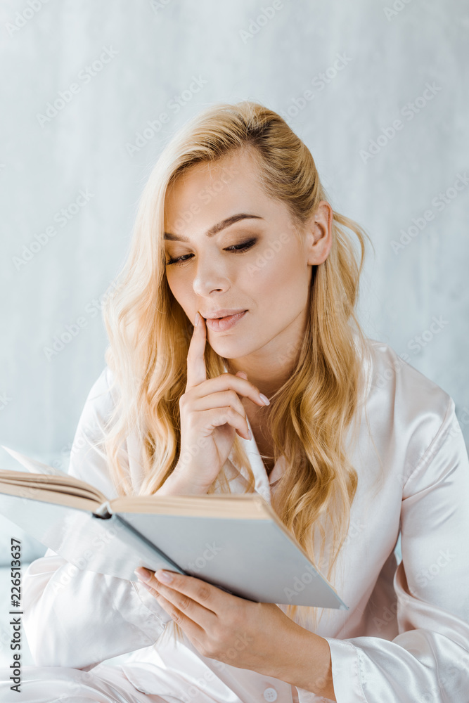 focused young blonde woman in pajamas reading book in bedroom focused young blonde woman in pajamas reading book in bedroom