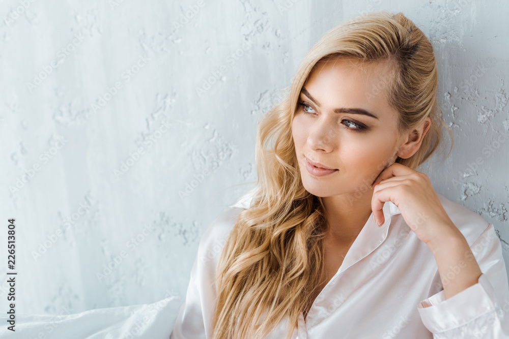 beautiful smiling young blonde woman in pajamas sitting on bed and looking away beautiful smiling young blonde woman in pajamas sitting on bed and looking away