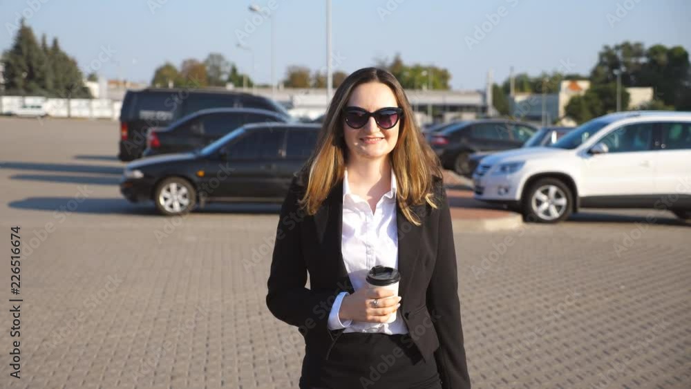 Young smiling businesswoman with cup of coffee walking near auto parking at sunny day. Happy business woman going to work. Positive confident girl being on his way to office. Slow motion Close up Young smiling businesswoman with cup of coffee walking near auto parking at sunny day. Happy business woman going to work. Positive confident girl being on his way to office. Slow motion Close up
