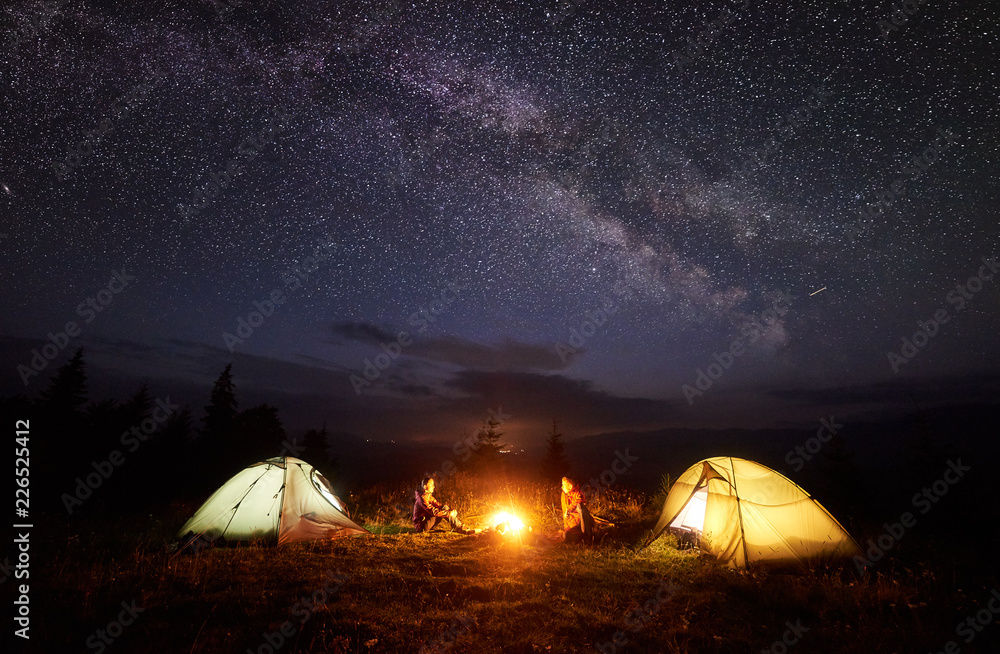 Camping in mountains at night. Bright bonfire burning between two hikers, boy and girl sitting opposite each other near illuminated tents under beautiful evening starry sky and Milky way - obrazy, fototapety, plakaty 
