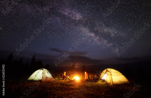 Camping in mountains at night. Bright bonfire burning between two hikers, boy and girl sitting opposite each other near illuminated tents under beautiful evening starry sky and Milky way