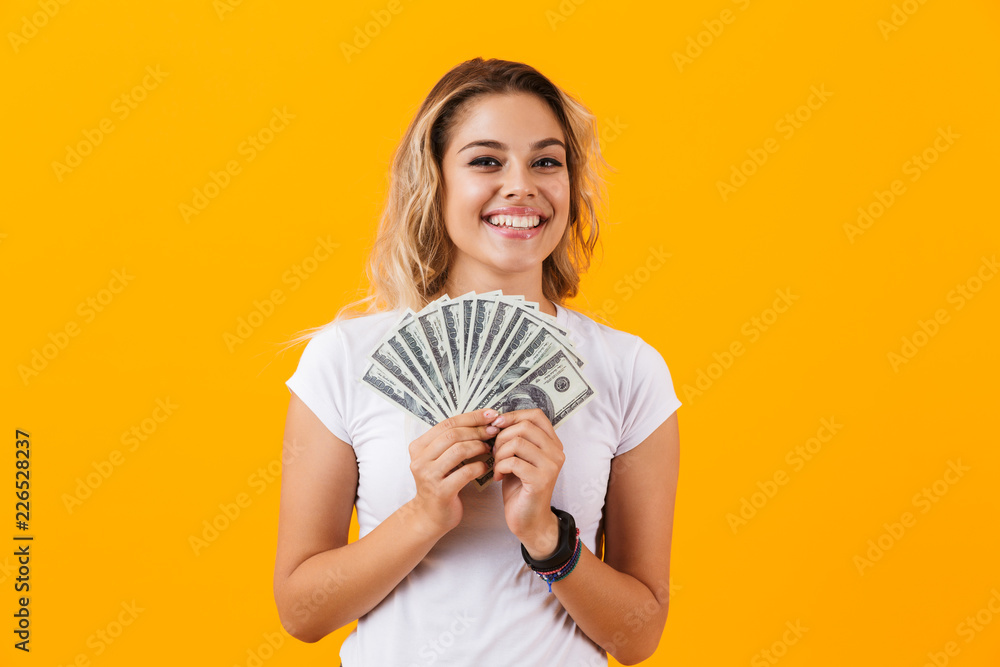 Photo of caucasian woman in basic clothing holding fan of dollar money, isolated over yellow background Photo of caucasian woman in basic clothing holding fan of dollar money, isolated over yellow background
