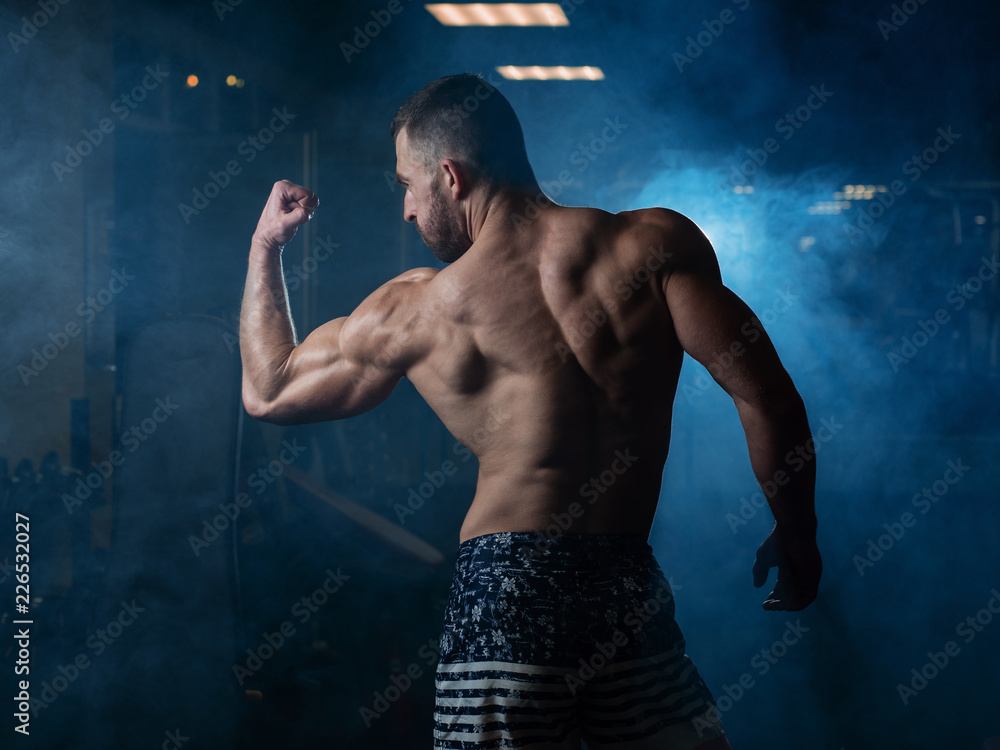 Fototapeta premium Athletic man with a muscular body poses in the gym, showing off his biceps and back. The concept of a healthy lifestyle