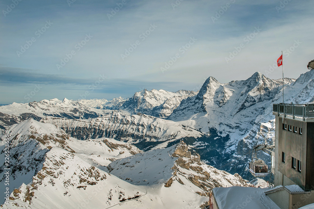 View of Alps in winter from Schilthorn peak. On the foreground the cableway from Birg to Schilthorn and part of station building with Switzerland flag on it/