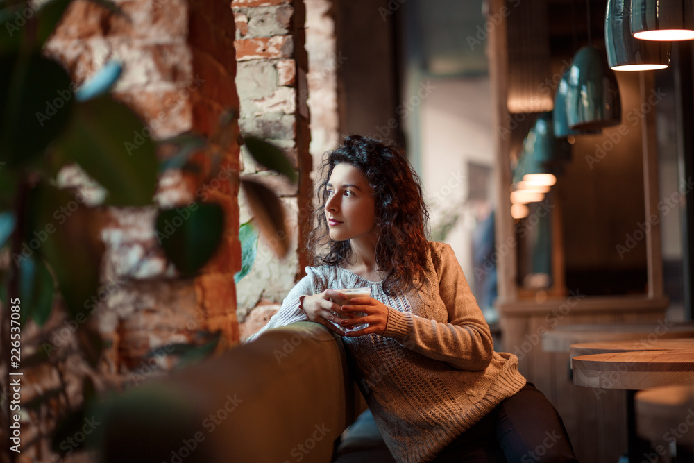 beautiful business girl drinking coffee from white cup in cozy cafe. sitting near window at a wooden table.Dressed in warm gray sweater