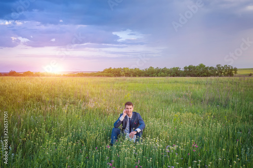 Businessman sitting in his suit on a green sunny field phoning from his mobile phone