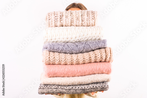 Close up of young woman holding huge stack of folded knitted warm pastel color sweaters, easy chic style, different knitting patterns. Light pink manicure. Background, copy space.