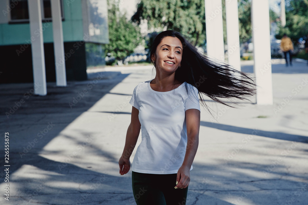 Young beautiful happy girl with long black hair, goes against the background of the city, hair develops from the wind. White T-shirt. Mockup.