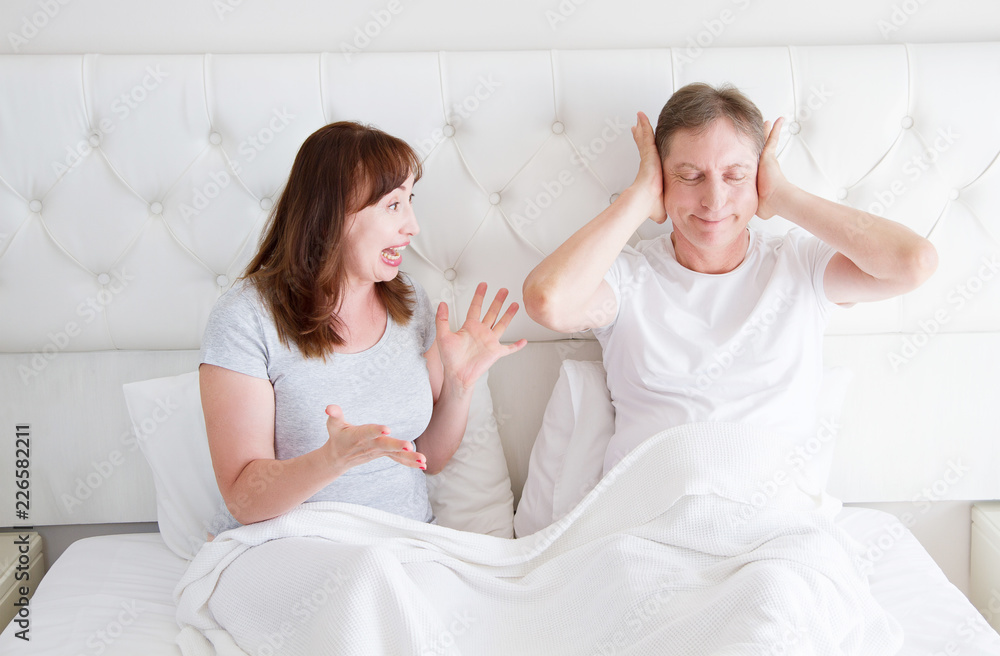 Caucasian middle age family couple angry shouting in bed. Conflict relationship concept. Husband cover ears by hands and wife trying make a dialogue. Selective focus