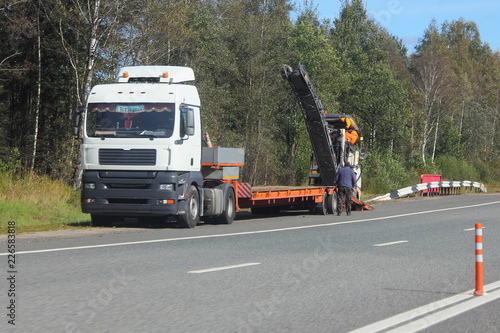 Low-frame european semi-trailer truck unloads asphalt remover summer day on the background of the roadside and green forest - transportation of special equipment for road repairs, transport logistics