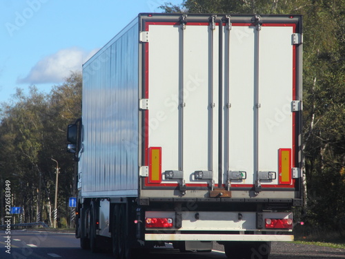 Heavy duty transport in logistics - white semi-trailer truck rides on asphalt highway in summer against green trees, road signs and blue sky, rear-side view