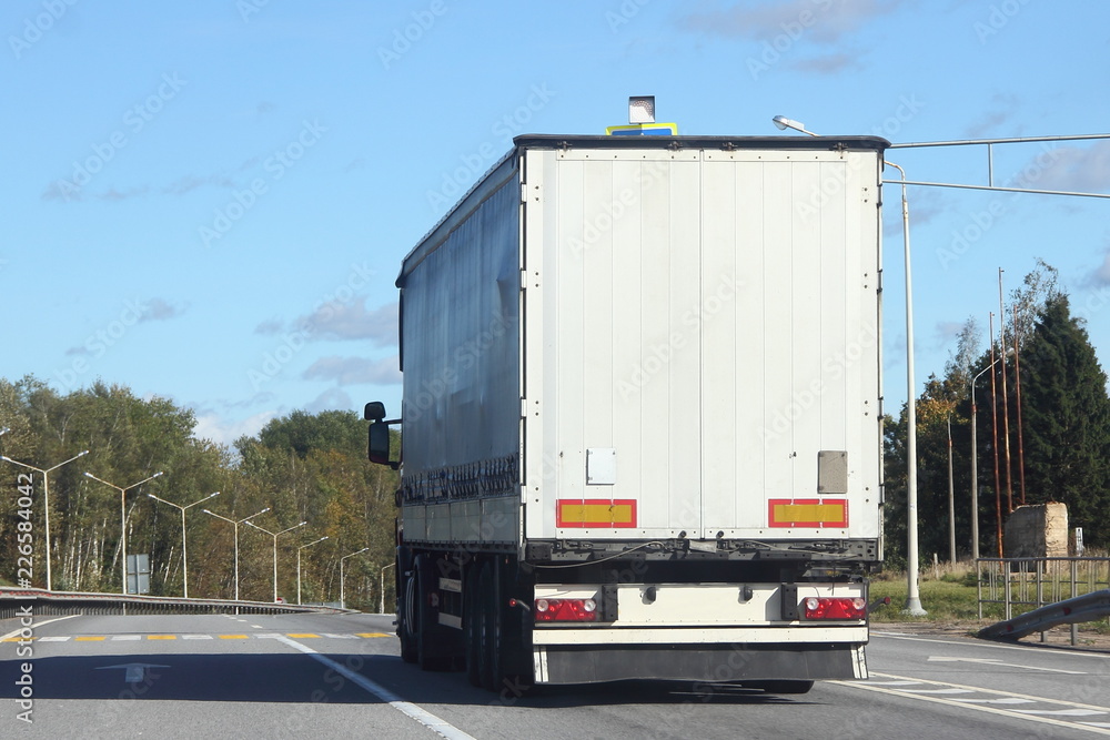 White truck with a semi-trailer go on a europe suburban highway road on a summer day against the blue sky and green forest, rear side view of the trailer - delivery, trade, transportation logistics White truck with a semi-trailer go on a europe suburban highway road on a summer day against the blue sky and green forest, rear side view of the trailer - delivery, trade, transportation logistics