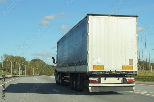 Beautiful white truck with a semi-trailer rides on turn of a suburban highway road in summer day against blue sky and green forest, side rear view of trailer - transportation, transport logistics