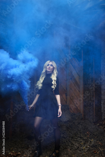 girl in a black short dress with white long wavy hair on a dark brown and black background with a smoke bomb and blue smoke