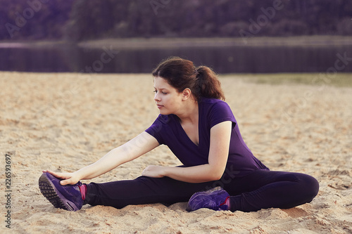 Pretty woman working out and stretching legs while sitting on sand beach near lake.