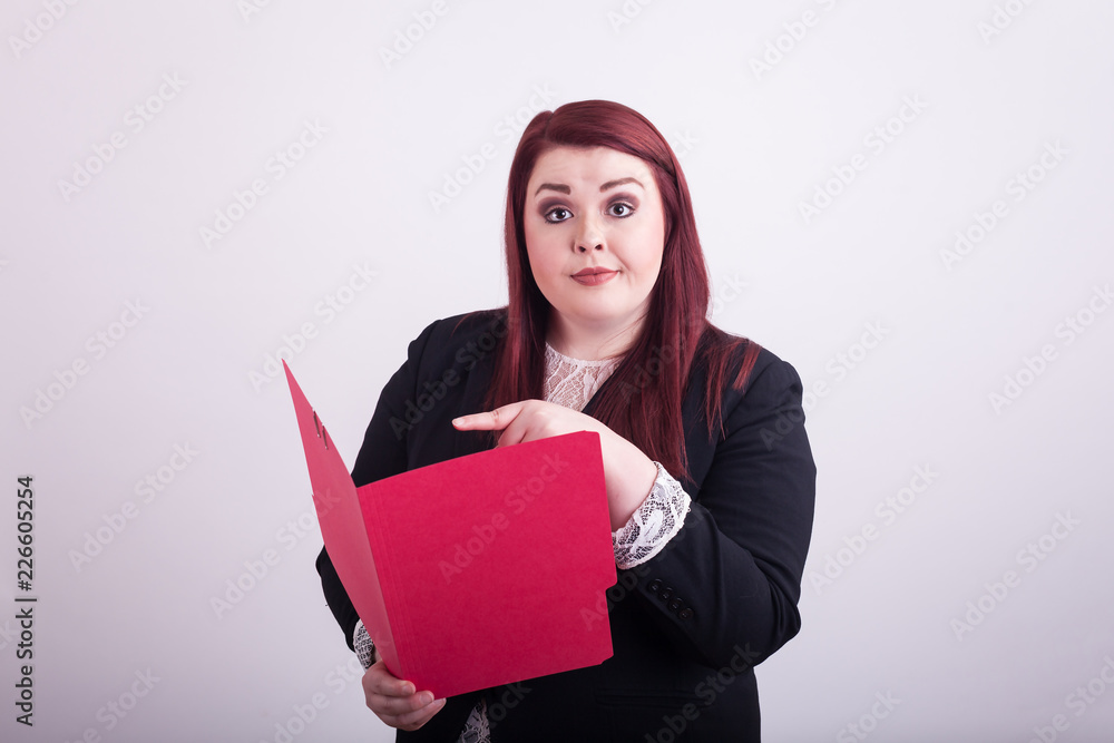 Young professional woman holding red folder pointing at folder looking at camera eyebrows raised Young professional woman holding red folder pointing at folder looking at camera eyebrows raised