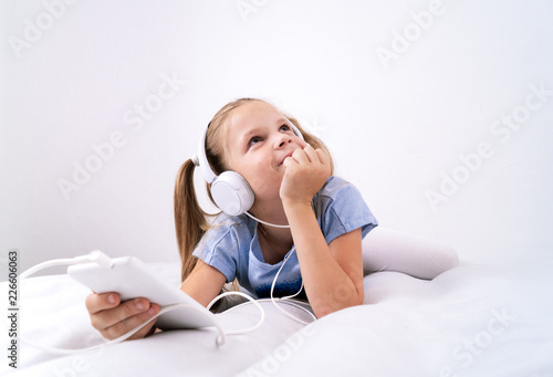 A girl in a blue t-shirt is lying on a white bed and listening to music in large white headphones using a player. Portrait.