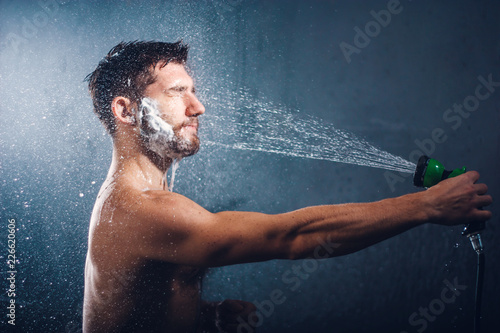 Headshot of a handsome bearded young man with eyes closed, holding a shower head and taking shower, with water splashes all over his face