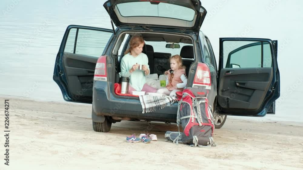 Beautiful young woman and her little daughter are sitting in the open trunk of a car on the bank of a river of the sea eating fruits and drinking tea from a thermos Beautiful young woman and her little daughter are sitting in the open trunk of a car on the bank of a river of the sea eating fruits and drinking tea from a thermos