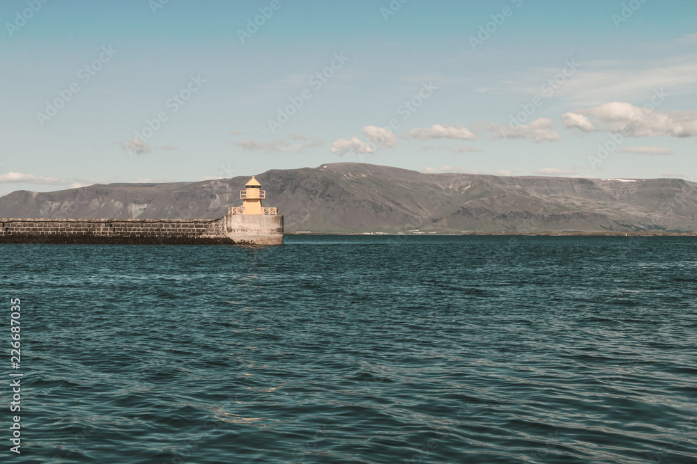 Yellow lighthouse in ocean bay against blue sky and mountains on horizon. Copy space. Yellow lighthouse in ocean bay against blue sky and mountains on horizon. Copy space.