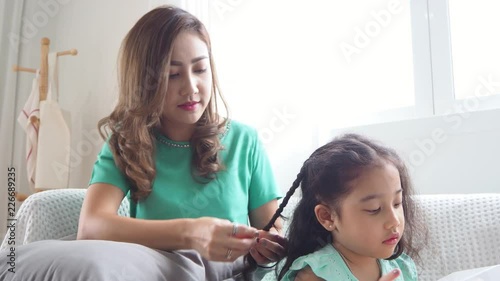 Beautiful asian mother braiding her daughter hair in living room at home
