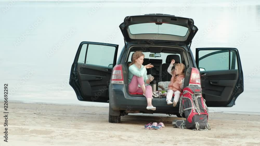 Beautiful young woman and her little daughter are sitting in the open trunk of a car on the bank of a river of the sea eating fruits and drinking tea from a thermos Beautiful young woman and her little daughter are sitting in the open trunk of a car on the bank of a river of the sea eating fruits and drinking tea from a thermos