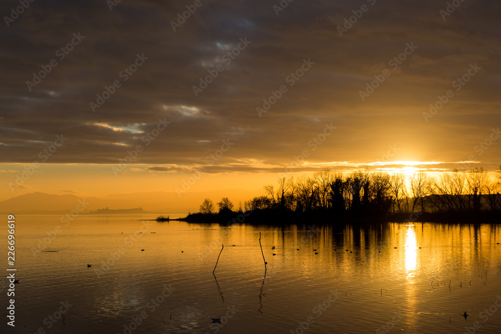 Beautiful view of Trasimeno lake at sunset, with orange tones, birds on water, Castiglione del Lago town on the background and sun reflecting on water