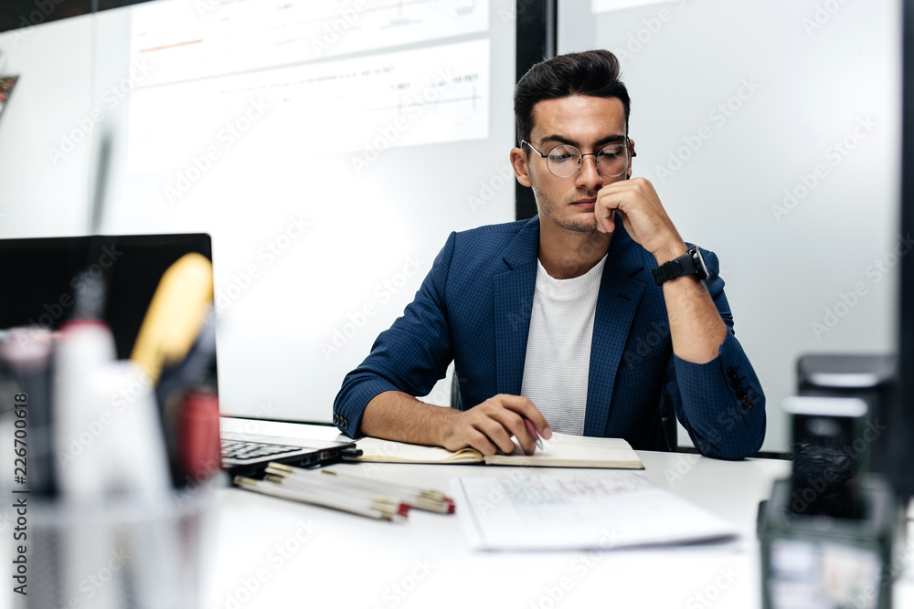 Dark-haired young architect in glasses and in a blue jacket is working with documents on the desk in the office Dark-haired young architect in glasses and in a blue jacket is working with documents on the desk in the office