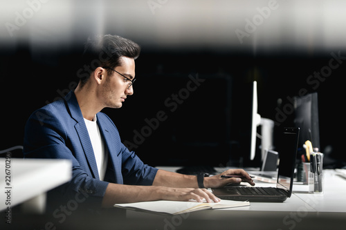 Architect in glasses dressed in blue checkered jacket makes notes in a notebook on the desk with laptop in the office