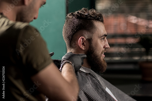 Brutal handsome man with beard sits at a barber shop. Barber shaves hairs at the neck © Leika production