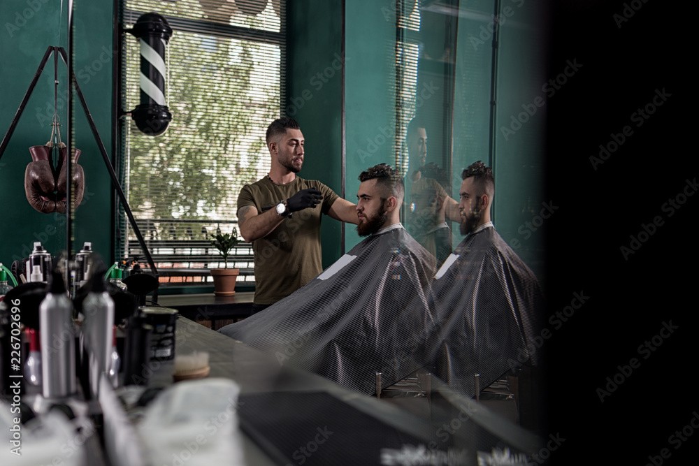 Man with a beard sits in the chair in front the mirror at a barber shop. Barber makes a hairstyle. Man with a beard sits in the chair in front the mirror at a barber shop. Barber makes a hairstyle.