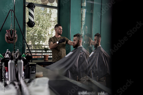 Man with a beard sits in the chair in front the mirror at a barber shop. Barber makes a hairstyle.