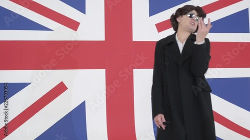 Wallpaper Mural Funny guy with black curly wig and sunglasses wearing long dark coat and white shirt is loudly and emotionally singing and gesturing with hands on camera in light studio with background of big UK flag Torontodigital.ca
