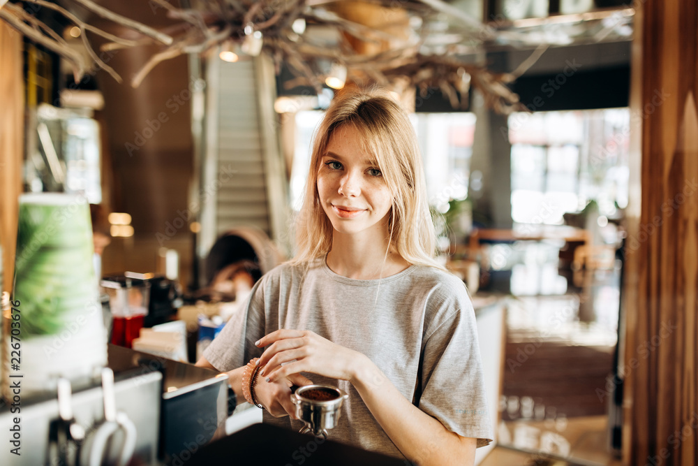 A pretty smiling slim girl,wearing casual clothes,holds some ground coffee and looks at the camera in a cozy coffee shop.