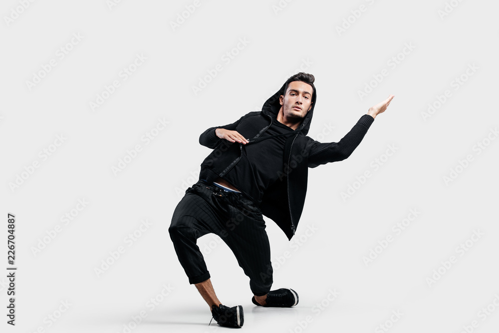 Dark-haired stylish dancer wearing black clothes and a hood makes stylized movements of street dance.