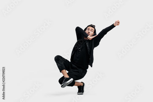 Dark-haired stylish dancer dressed in a black clothes makes stylized movements of street dance. He is sitting on one leg and put his hand up