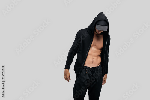 Handsome young dancer dressed in a sweatshirt on a naked torso with a hood on the cap and black pants stands on a white background