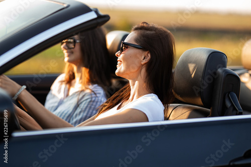 Two beautiful young women in sunglasses are sitting in a black cabriolet and smiling on a sunny day. © Leika production
