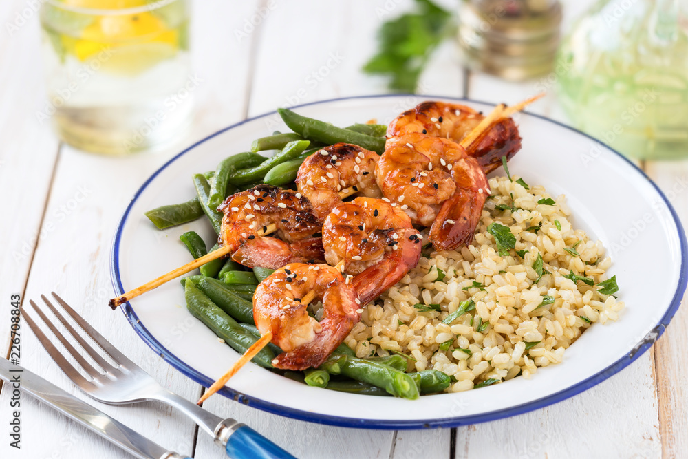 Buddha bowl with brown rice, green beans and shrimp skewers on white wooden table
