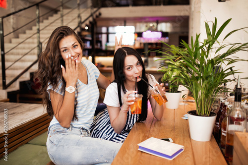 Two pretty youthful smiling girls,dressed in casual outfit,sit next to each other and look at the camera in a cozy coffee shop.
