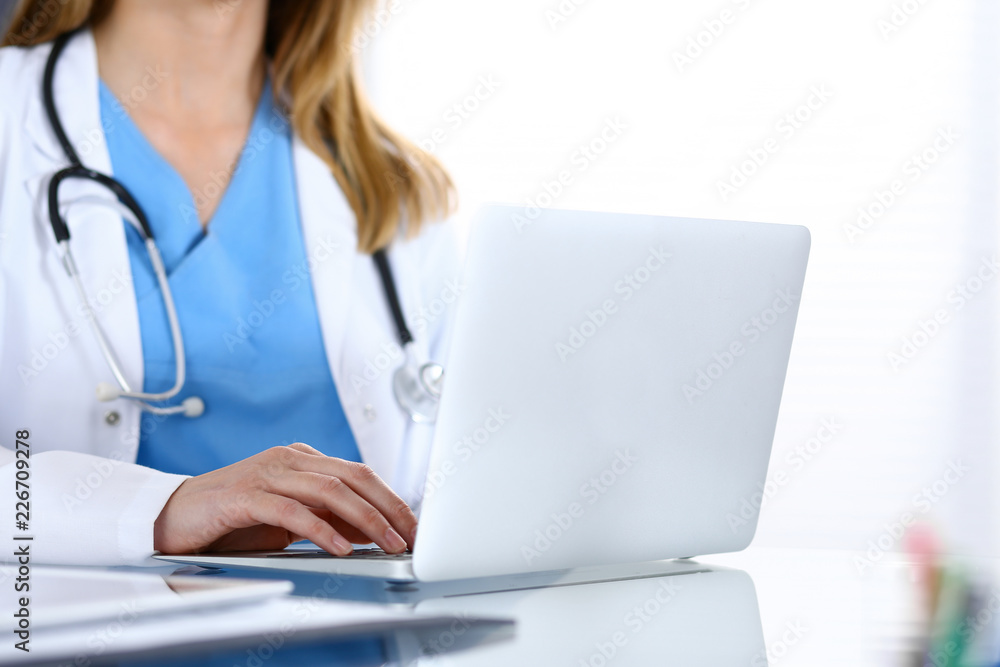 Doctor typing on laptop computer while sitting at the glass desk in hospital office. Physician at work. Medicine and healthcare concept Doctor typing on laptop computer while sitting at the glass desk in hospital office. Physician at work. Medicine and healthcare concept