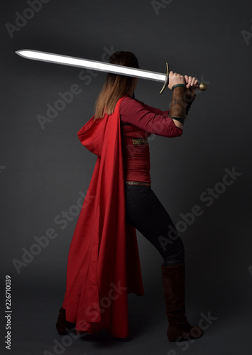 full length portrait of brunette girl wearing red medieval costume and cloak. standing pose with back to the camera, holding a sword on grey studio background.