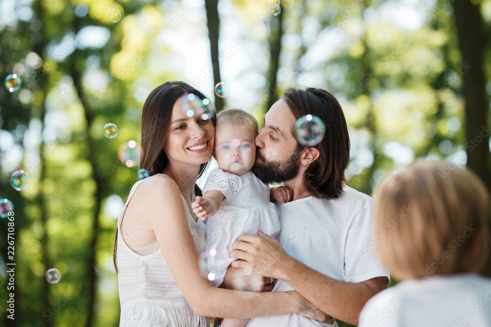 Smiling parents dressed in the white clothes are holding little daughter in the arms and looking at the soap bubbles. Smiling parents dressed in the white clothes are holding little daughter in the arms and looking at the soap bubbles.