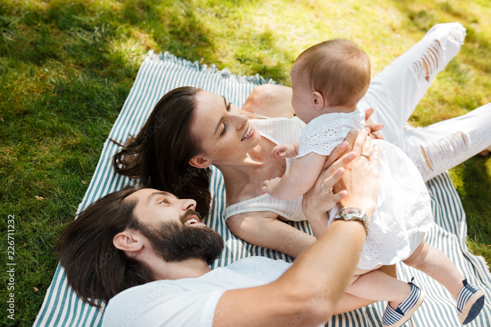 Young dark-haired mother and father dressed in white clothes are lying on a striped plaid on a law holding in the arms little daughter. Young dark-haired mother and father dressed in white clothes are lying on a striped plaid on a law holding in the arms little daughter.