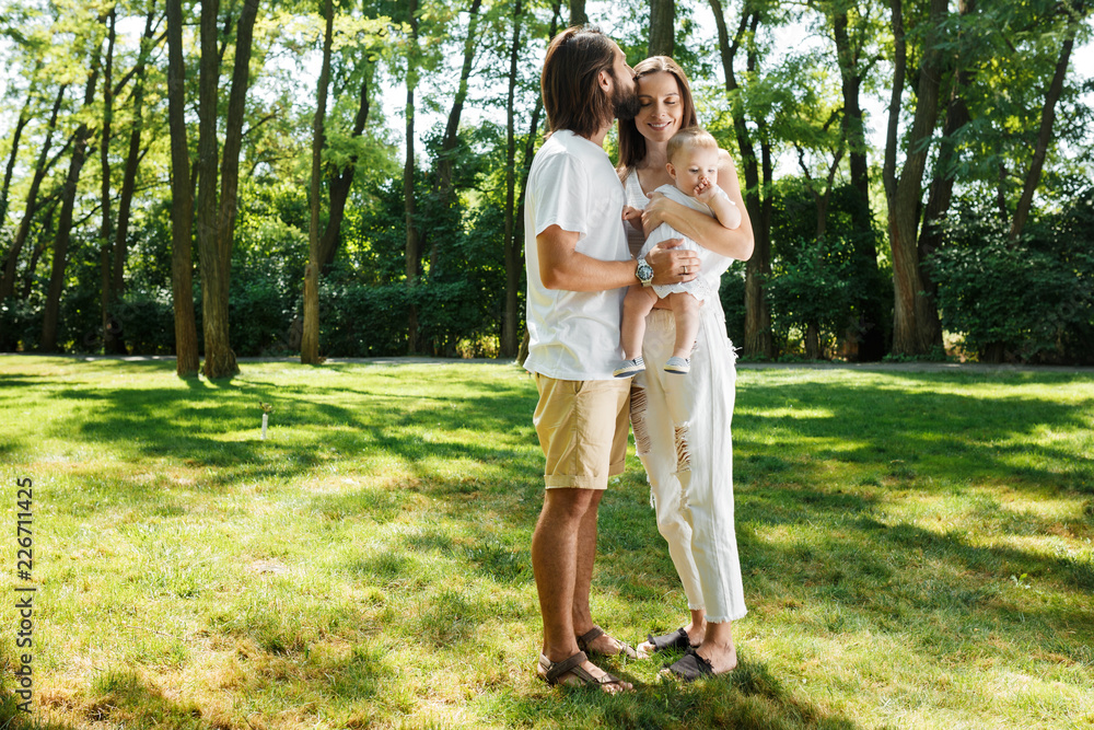 Dark-haired father kisses his beautiful wife holding a small daughter in a white dress in a park on a sunny day. Dark-haired father kisses his beautiful wife holding a small daughter in a white dress in a park on a sunny day.